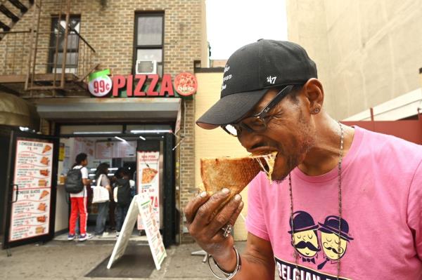 A man eating a pizza slice in front of a 99-cent pizza shop 