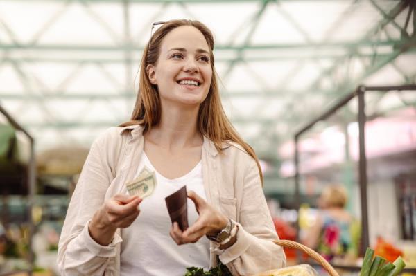 A smiling young woman is holding a basket full of groceries and counting money.