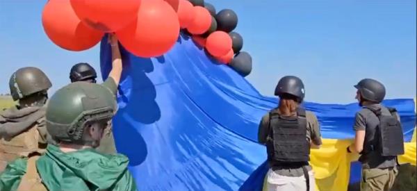 Red and black balloons begin to rise into the sky, with a giant blue and yellow Ukranian flag attached, as soldiers wearing helmets assist. 
