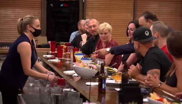 jon taffer stands at a bar full of people and points to a drink while a bartender in a facemask prep behind the bar