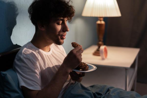 A stressed young man in a white t-shirt sitting in bed at night, eating a coo<em></em>kie. a white nightstand with a lamp and a clock on top are next to the bed
