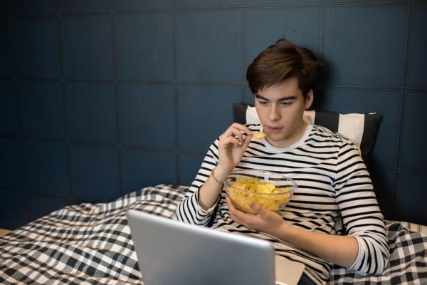 Teenage boy using laptop in bed and eating chips while watching movies at home.