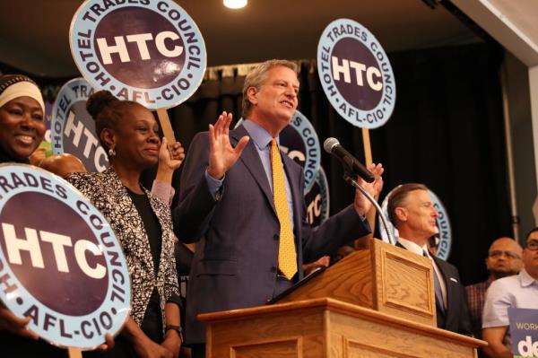 Bill de Blasio standing at a podium at a rally as the Hotel and Motel Trades Council unio<em></em>n endorses him, with Chirlane McCray in the audience.