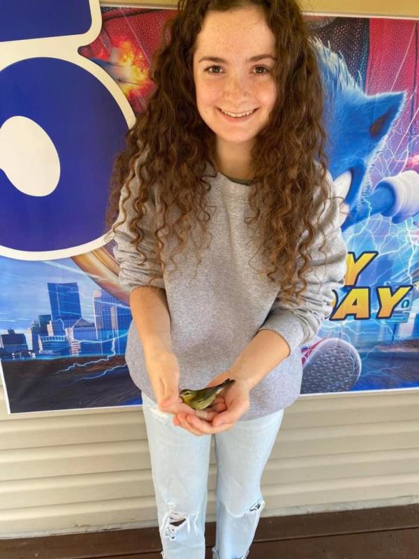 Autumn McClure smiling, holding a small bird in her hands, seen wearing gray sweatshirt and jeans standing in font of a colorful sign.