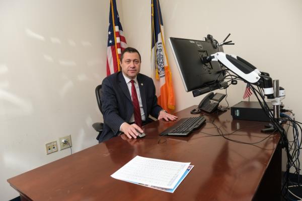 A man in a dark pinstripe suit and a red tie sits at a desk in front of a large computer mo<em></em>nitor with his hand on the mouse with the American flag and New York City flag behind him.