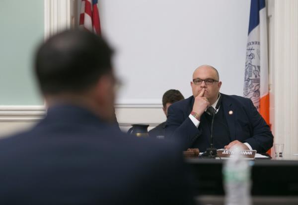 A man wearing a navy suit with a microphone in front of him presses his finger to his lips as he sits between an American flag and a New York City flag.