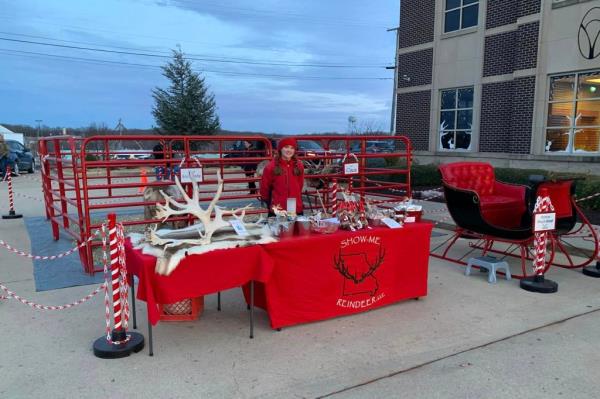One of the Shulls stands at a table covered with a red cloth with reindeer antlers and other items on top, in front of the pen used to keep the reindeer.