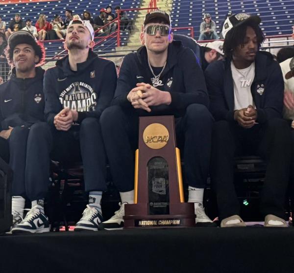 UConn basketball players Jaylin Stewart, Alex Karaban, Do<em></em>novan Clingan, and Tristen Newton sitting on stage with the natio<em></em>nal champio<em></em>nship trophy