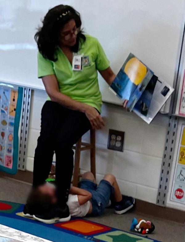 A stunning photo snapped by a teacher's aide in the room shows Otero, 59, sitting in a chair and reading a book to the class as the boy lies on his back beneath her in the leg grip.