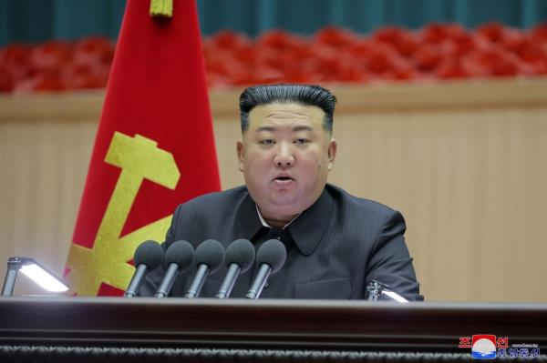 North Korean leader Kim John Un sits in front of a bank of five micropho<em></em>nes with the North Korean flag on a pole behind his right shoulder.