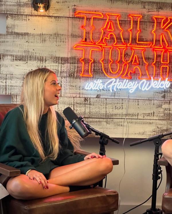 Haliey Welch sitting in a chair with her legs crossed while speaking into a microphone on her podcast 
