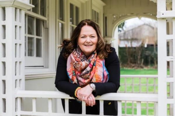 Alison wearing a colourful scarf and black jacket is leaning on a patio fence, looking to camera and smiling